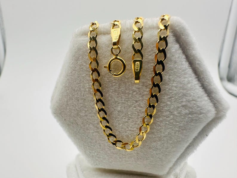 May include: A gold-colored curb chain necklace with a lobster clasp and a separate chain segment with a spring ring clasp. The chain is displayed on a white, textured surface. The links are flat and oval.