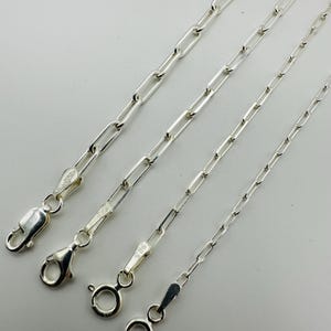 May include: Four silver paperclip chain necklaces of varying lengths are displayed against a white background. Each chain features a different clasp style. The chains are made of shiny, polished metal, reflecting the light.
