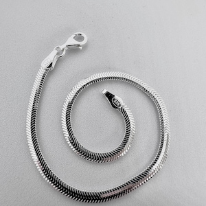 May include: A silver-colored bracelet with a herringbone chain design. The bracelet is made of a shiny, reflective metal and features a lobster clasp closure. The bracelet is coiled in a spiral shape.