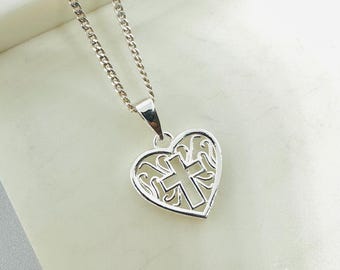 Può includere: Una collana color argento con un ciondolo a forma di cuore con un design a croce. Il ciondolo ha un design traforato. La catena è a maglia curb. Lo sfondo è di colore chiaro e neutro.