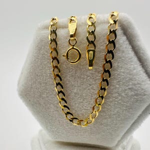 May include: A gold-colored curb chain necklace with a lobster clasp. The chain is displayed on a white, textured surface. The necklace is made of interlocking oval links. The clasp and chain links are visible.