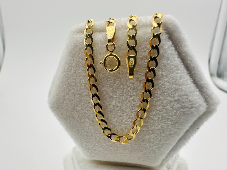 May include: A gold-colored curb chain necklace with a lobster clasp. The chain is displayed on a white, textured surface. The necklace is made of interlocking oval links. The clasp and chain links are visible.