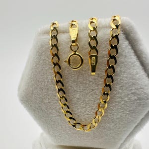 May include: A gold-colored curb chain necklace with a lobster clasp and a separate chain segment. The necklace is displayed on a white, textured surface. The chain links are oval-shaped and closely connected.