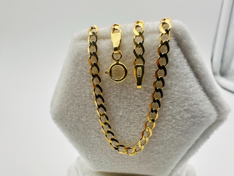 May include: A gold-colored curb chain necklace with a lobster clasp and a separate chain segment. The necklace is displayed on a white, textured surface. The chain links are oval-shaped and closely connected.