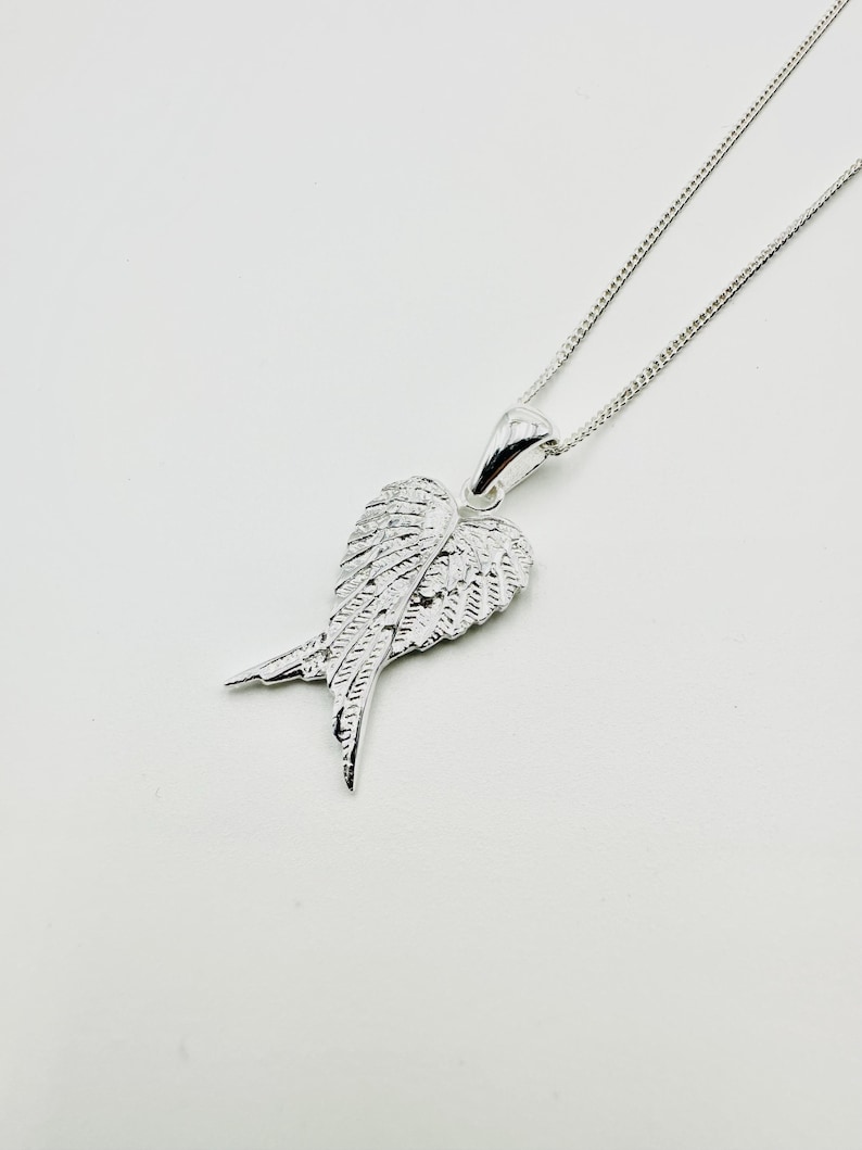 Puede incluir: Collar con colgante plateado en forma de ala. El colgante, con detalles de plumas, cuelga de una fina cadena. El collar se presenta sobre un fondo blanco. El dise&ntilde;o de ala es un s&iacute;mbolo popular de libertad y protecci&oacute;n.