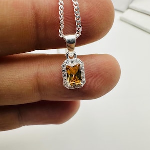 May include: Silver necklace with a rectangular, amber-colored gemstone pendant. The pendant is surrounded by small, clear stones. The chain is a curb style. The jewelry is displayed against a neutral background.