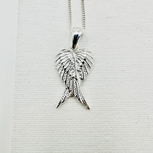 Puede incluir: Un collar con colgante plateado con un dise&ntilde;o de ala detallado. El colgante, con forma de coraz&oacute;n, presenta intrincados detalles de plumas. La cadena fina y el colgante se exhiben sobre un fondo blanco.