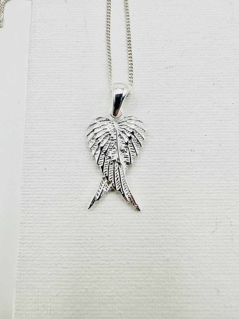 Puede incluir: Un collar con colgante plateado con un dise&ntilde;o de ala detallado. El colgante, con forma de coraz&oacute;n, presenta intrincados detalles de plumas. La cadena fina y el colgante se exhiben sobre un fondo blanco.