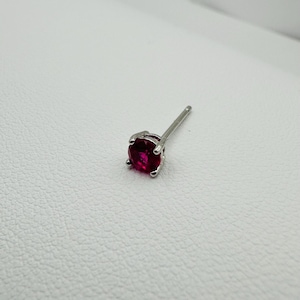 Może przedstawiać: Pojedynczy srebrny kolczyk z okrągłym, głęboko czerwonym kamieniem szlachetnym. Kamień jest utrzymywany na miejscu przez cztery pazury. Sztyft kolczyka jest widoczny i wystaje z tyłu. Kolczyk na białym tle.