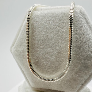May include: A silver snake chain necklace displayed on a white, textured surface. The necklace has a smooth, flexible design, with interlocking links. The chain is approximately 45 cm long. The chain is a bright, reflective silver.