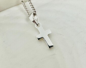May include: A silver-colored cross pendant with a matching chain. The cross is a simple, polished design with rounded edges. The chain is a curb style, and the pendant hangs from a small loop at the top of the cross. The jewelry is displayed against a light background.