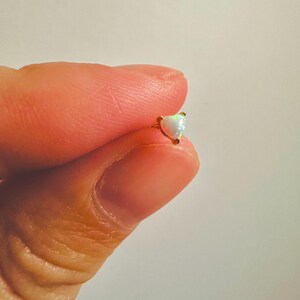 May include: A tiny heart-shaped earring with a gold setting and a white opal stone. The earring is held between two fingers against a plain background. The opal has a subtle iridescent quality.