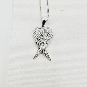 Puede incluir: Un collar con colgante plateado con un dise&ntilde;o detallado de alas. El colgante cuelga de una fina cadena. Las alas est&aacute;n intrincadamente detalladas, con cada pluma claramente definida. El collar se muestra sobre un fondo blanco.