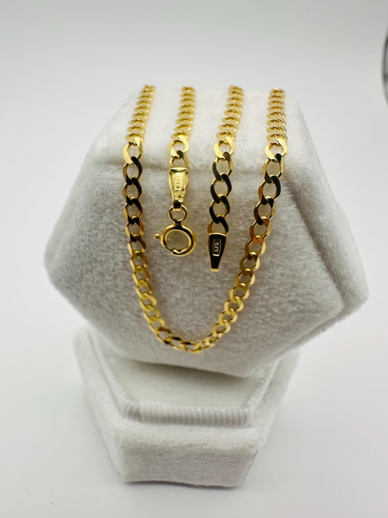 May include: A gold chain necklace with a curb link design, displayed on a white velvet jewelry box. The necklace is a bright, shiny gold color, with a lobster clasp closure. The chain is approximately 18 inches long.