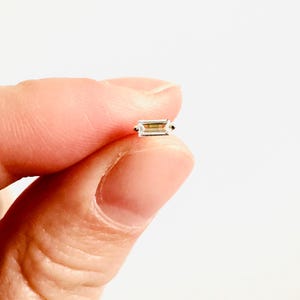 Puede incluir: Una pequeña gema rectangular con una apariencia clara y translúcida. La gema se sostiene entre los dedos sobre un fondo blanco, mostrando su forma y claridad. Es probable que la gema sea una pieza de joyería.