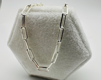 May include: A silver chain necklace with elongated rectangular links. The necklace is displayed on a soft, light grey, hexagonal surface against a white background. The chain is made of a shiny, reflective metal.