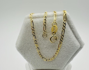 May include: A gold-colored Figaro chain necklace with an oval and elongated link design. The necklace is displayed on a gray, textured surface. The clasp and extension chain are also visible.