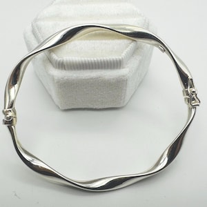 May include: A silver-toned, twisted bangle bracelet. The bracelet has a continuous, flowing design with a clasp closure. The bracelet is displayed on a white, textured surface.