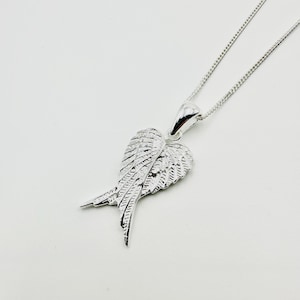 May include: A silver pendant necklace with a wing-shaped design. The pendant, with detailed feathering, hangs from a fine chain. The necklace is presented on a white backdrop. The wing pendant's intricate detailing gives it a textured appearance.
