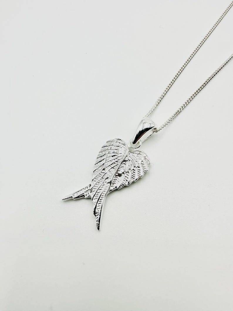 Puede incluir: Collar con colgante plateado en forma de ala. El colgante, con detalles de plumas, cuelga de una fina cadena. El collar se presenta sobre un fondo blanco. Los intrincados detalles del ala le dan una apariencia texturizada.