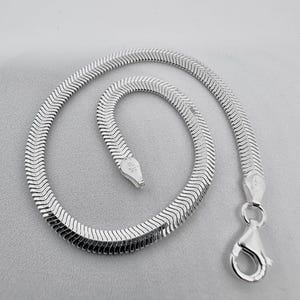 May include: A silver herringbone chain bracelet with a lobster clasp. The bracelet is composed of small, flat, interlocking metal pieces, creating a flexible, textured surface. The clasp is attached to the end of the chain.