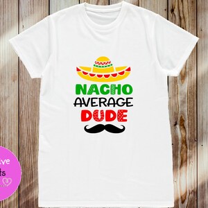 Peut inclure: T-shirt blanc avec un motif graphique coloré. Le motif représente un sombrero, les mots "NACHO AVERAGE DUDE" en vert et rouge, et une moustache noire. Le t-shirt est présenté sur un fond en bois.