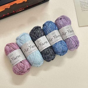 May include: Five skeins of Air Yarn in various colors: light pink, light blue, dark grey, blue, and purple. Each skein has a white label with the text "Air Yarn" and "Weaving a better life". The yarn is neatly arranged.
