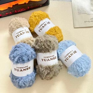Puede incluir: Un conjunto de ovillos de lana esponjosa en varios colores, incluyendo beige, amarillo, gris y azul. Cada ovillo tiene una etiqueta blanca con el texto "YUSHANG YUANSE". La lana parece suave y texturizada.