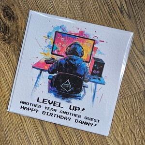 Peut inclure: Une carte d'anniversaire avec une illustration color&eacute;e d'une personne assise devant un &eacute;cran d'ordinateur. Le texte sur la carte est "Level Up! Another year another quest Happy Birthday Danny!"