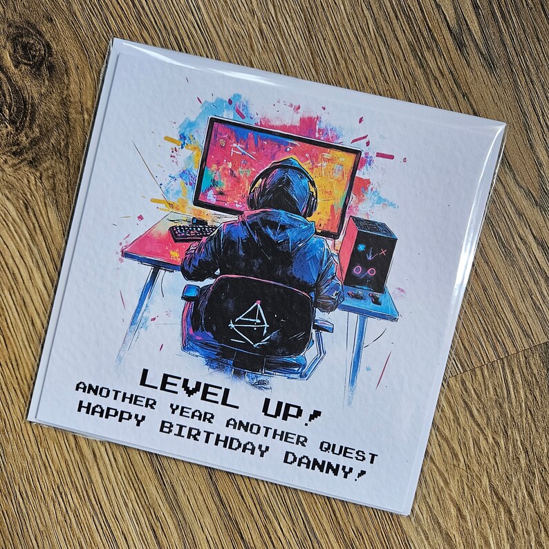 Peut inclure: Une carte d'anniversaire avec une illustration color&eacute;e d'une personne assise devant un &eacute;cran d'ordinateur. Le texte sur la carte est "Level Up! Another year another quest Happy Birthday Danny!"
