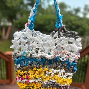 May include: A crocheted tote bag made from recycled plastic bags. The bag has a blue handle and is made with white, blue, yellow, and pink plastic.