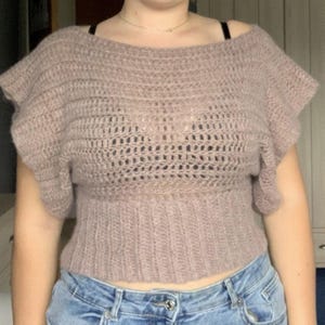 Puede incluir: Un top de ganchillo malva con un amplio escote barco, mangas cortas con volantes y un cuerpo de punto abierto y texturizado que termina en una cintura acanalada y ajustada. La prenda está confeccionada con hilo suave y difuso.
