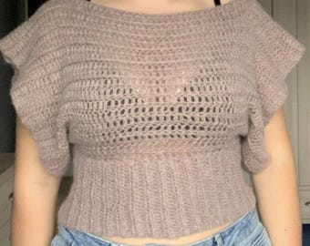 May include: A mauve crochet top featuring a wide boat neckline, short ruffled sleeves, and a textured, open-knit bodice that transitions into a ribbed, fitted waistband. The garment is crafted with a soft, fuzzy yarn texture.