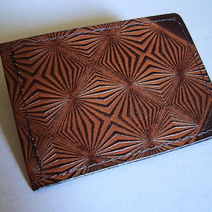 May include: A brown leather wallet with a geometric starburst pattern. The wallet is rectangular and features a repeating design of dark brown lines radiating from central points. The edges are stitched with light-colored thread.