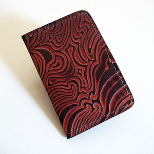 May include: A rectangular card holder featuring a deep red and black marbled pattern with a textured, embossed surface. The edges are finished with visible dark stitching, creating a structured appearance for this small, flat accessory.