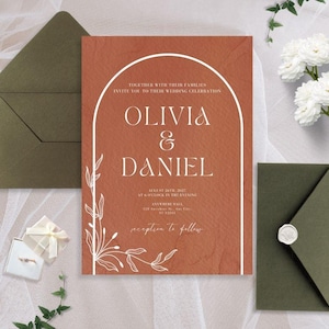 Puede incluir: Una invitación de boda con fondo terracota y texto blanco. La invitación presenta un diseño de arco simple con un motivo floral y los nombres "Olivia & Daniel". El texto dice "Junto con sus familias, les invitan a su celebración de boda".