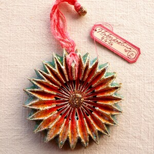 May include: A handmade, starburst-shaped ornament with a red, orange, and gold color scheme. The ornament has a teal border and a pink string for hanging. A small tag reads "Haberdashing co. 4.6.26".