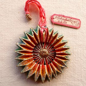 May include: A handmade, starburst-shaped ornament with a red and gold glittered loop for hanging. The ornament features a layered design with orange, green, and gold accents. A small tag with handwritten text is attached.
