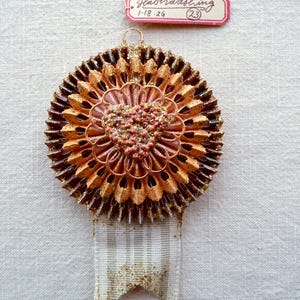 May include: A handcrafted, circular ornament with a layered, textured design in shades of gold, brown, and pink. The center features a heart-shaped embellishment with small, red and gold beads. A ribbon with gold glitter accents hangs below. The tag reads "Haberdashing 1-18-26 (23)".