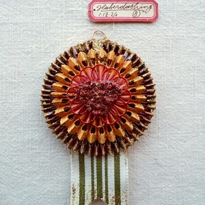 May include: A vintage, handcrafted ornament with a layered, flower-like design. The ornament features shades of brown, gold, and red, with a striped ribbon and a small tag that reads "Haberdashing".