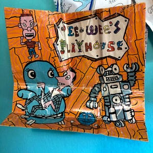 May include: A colorful illustration featuring the text "Pee-Wee's Playhouse" in a playful font. The artwork includes cartoon characters, a robot, and a blue creature, all set against an orange background with a wood-grain pattern.