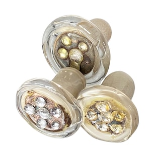 May include: Three clear glass bottle stoppers with decorative metallic accents. Each stopper has a rounded, light-colored cork and a clear glass top filled with silver and gold-colored embellishments. The stoppers are arranged in a cluster, showcasing their unique design.
