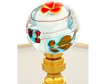 May include: A glass lamp finial with a gold base. The glass is clear with colorful flowers and swirls inside.