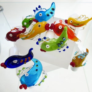 Puede incluir: Colección de figuras de peces de vidrio coloridas y hechas a mano. Cada pez presenta un diseño único con tonos vibrantes de azul, verde, rojo y amarillo. Los peces se exhiben sobre una superficie blanca, mostrando sus intrincados detalles y formas lúdicas.