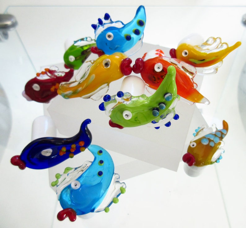 May include: A collection of colorful, handcrafted glass fish figurines. Each fish features a unique design with vibrant hues of blue, green, red, and yellow. The fish are displayed on a white surface, showcasing their intricate details and playful shapes.