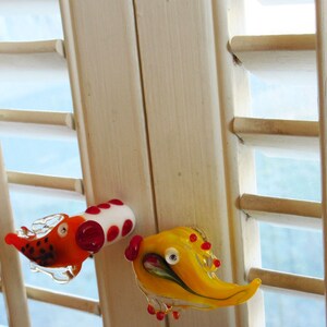 May include: Two whimsical glass figurines. One is orange and white with red accents, resembling a dog. The other is yellow with red accents, possibly a bird. Both are transparent and appear to be handmade, with a playful design.