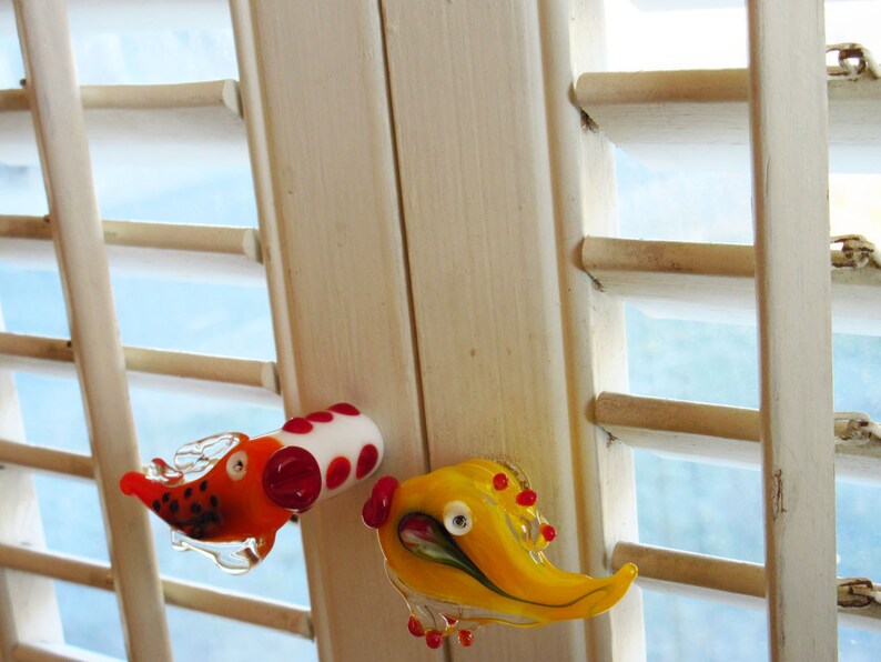 May include: Two whimsical glass figurines. One is orange and white with red accents, resembling a dog. The other is yellow with red accents, possibly a bird. Both are transparent and appear to be handmade, with a playful design.