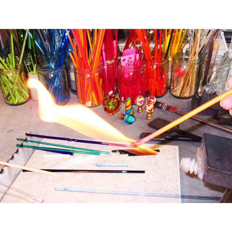 May include: A glassblowing scene with a flame torch heating a glass rod. Jars filled with colorful glass rods are in the background. Several finished glass art pieces are on the table, including small decorative globes and a colorful spiral.