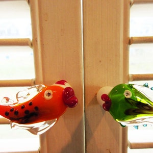May include: Two colorful glass fish door knobs. One is orange with black spots and a red accent, the other is green with blue accents. Both have white and red details and are mounted on a white surface.