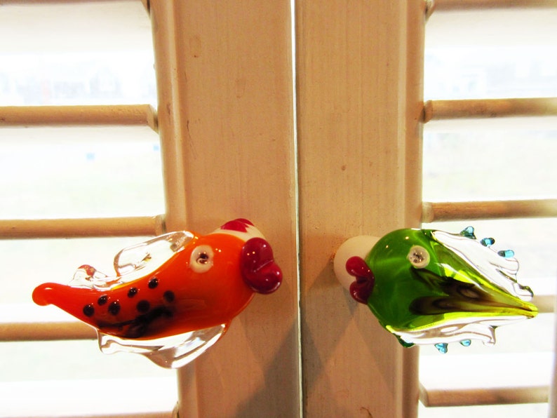 May include: Two colorful glass fish door knobs. One is orange with black spots and a red accent, the other is green with blue accents. Both have white and red details and are mounted on a white surface.